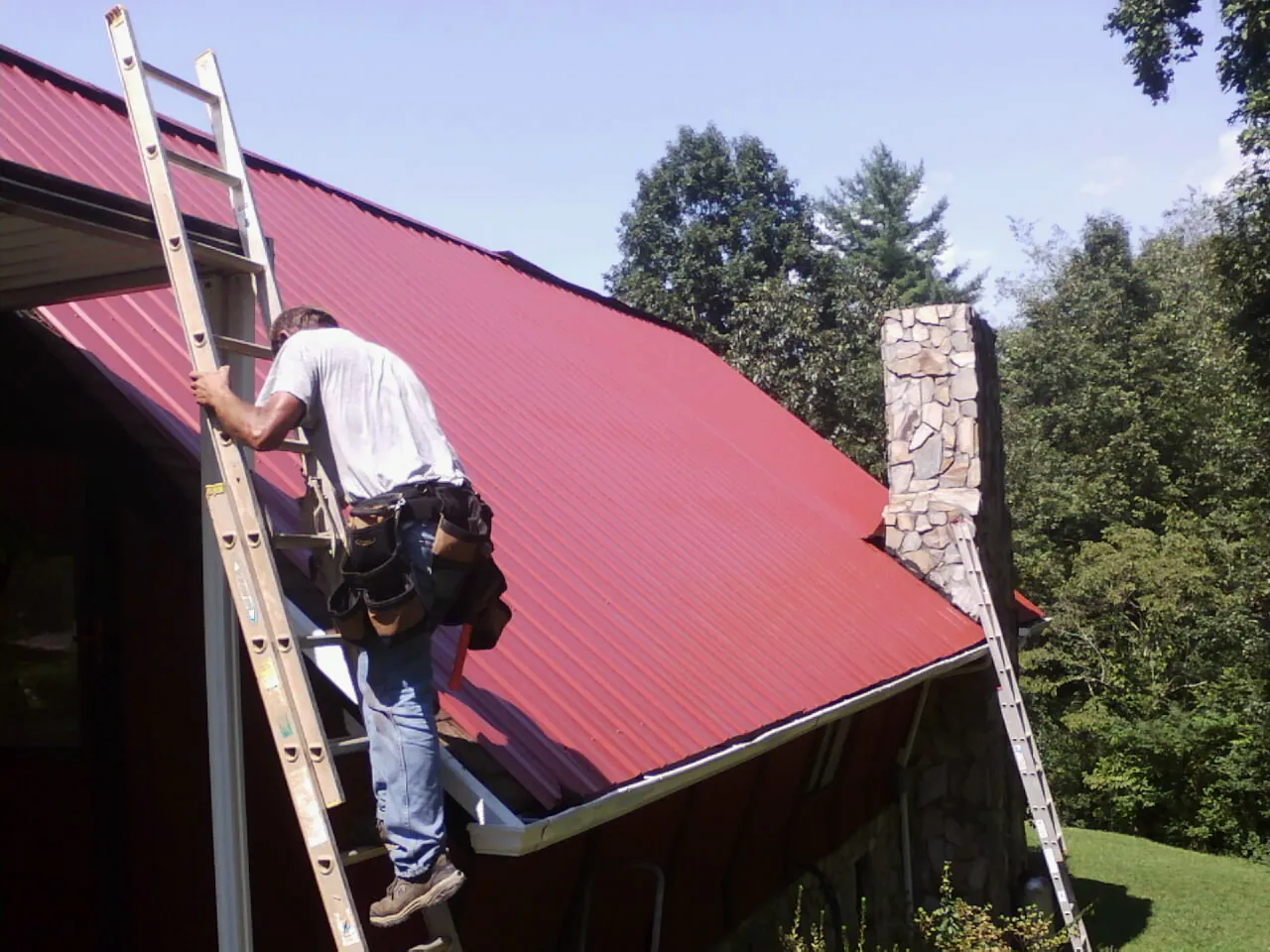 Roofer on a ladder beside a red metal roof and stone chimney — Soffit & Fascia Repair by Roofing Brothers in Bronxville